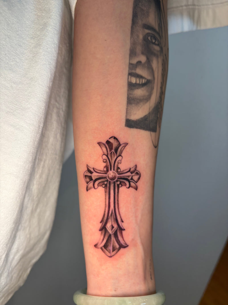A close-up of a person's forearm featuring a detailed black and gray gothic cross tattoo, with a partial grayscale portrait tattoo above it.