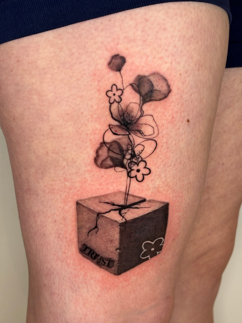 A close-up of a thigh showing a black and gray tattoo of a flower blooming from a cube with the word 'TRUST' on it.