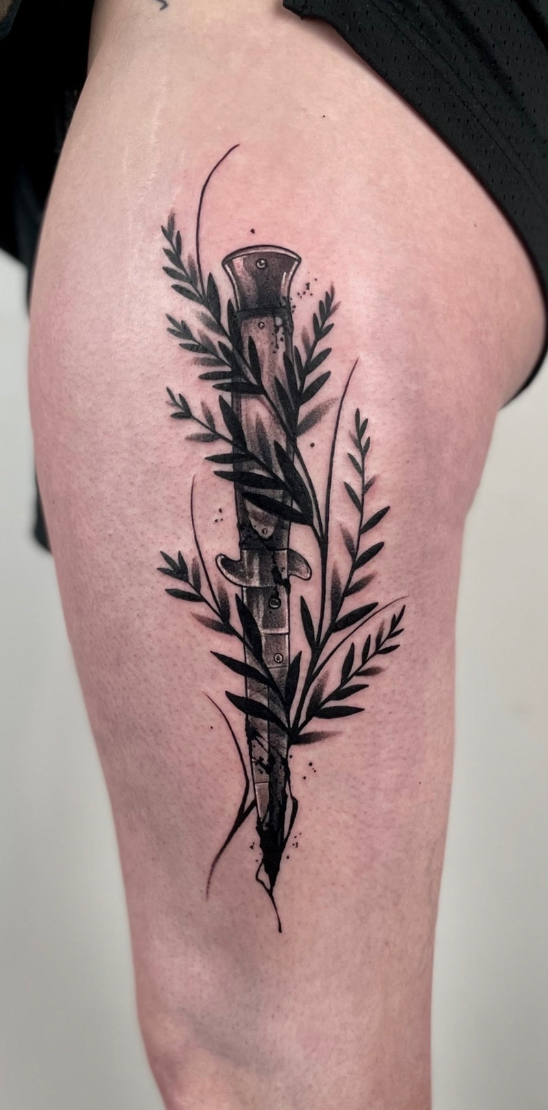 A black and gray tattoo of a dagger surrounded by leaves on a person's upper thigh,
