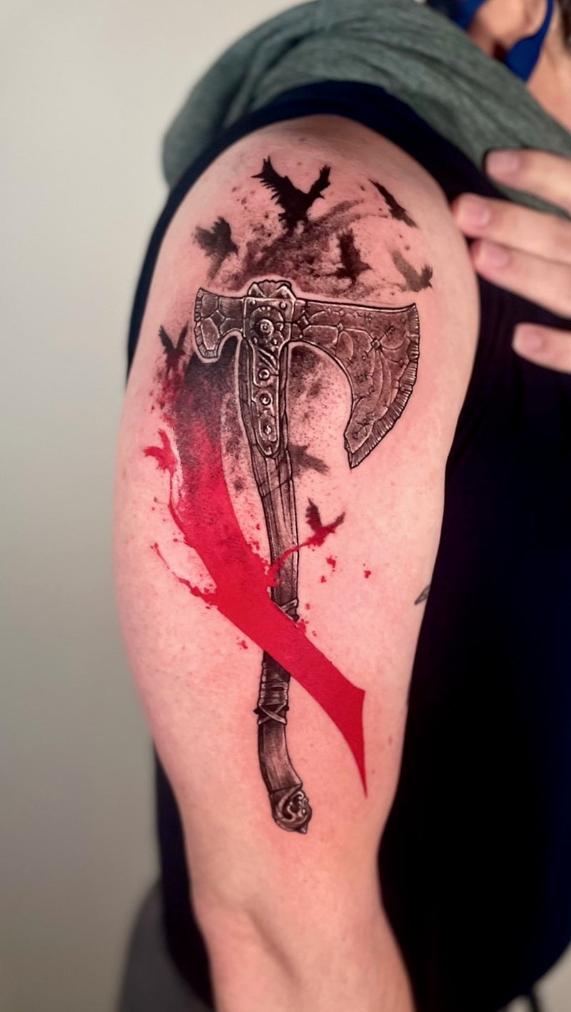 A close-up of a person's upper arm featuring a detailed black and gray tattoo of a Viking-style battle axe with a bold red slash design and black bird silhouettes on the shoulder.
