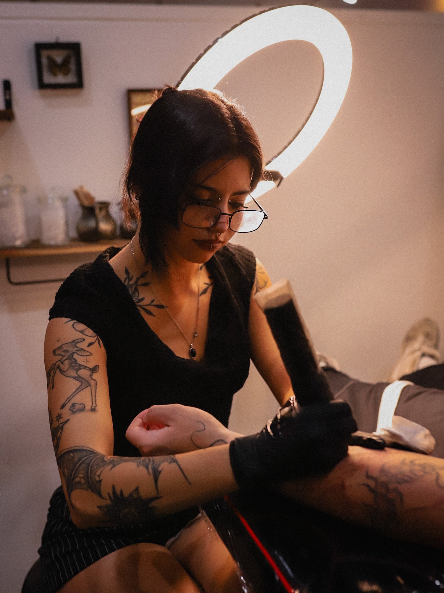 A tattoo artist with arm tattoos and glasses, wearing a black top and gloves, is shown tattooing a client's arm in a studio with a large ring light.