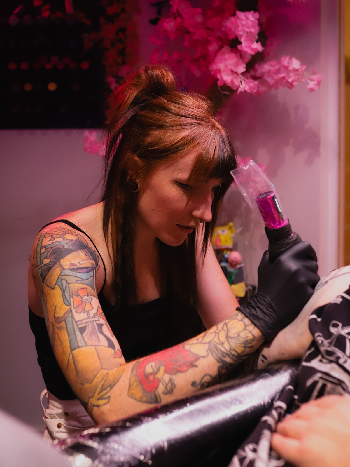 A tattoo artist with colorful arm tattoos wears a black tank top and black gloves while holding a tattoo pen machine. The scene is lit with pink ambient lighting, with cherry blossom decorations in the background.