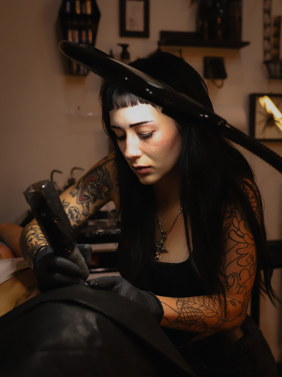 A tattoo artist with arm tattoos wearing black gloves and holding a tattoo machine over a client's shoulder in a dimly lit tattoo studio.