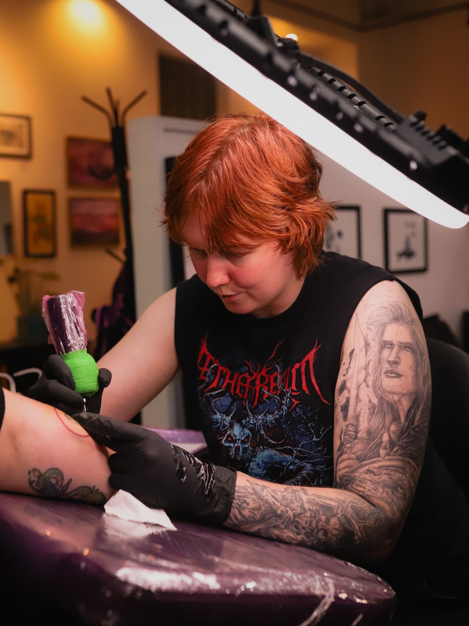 A tattoo artist with red hair and arm tattoos works on a client's arm, wearing a black sleeveless shirt with a red and blue graphic and black gloves. A tattoo lamp is visible overhead in a tattoo studio.