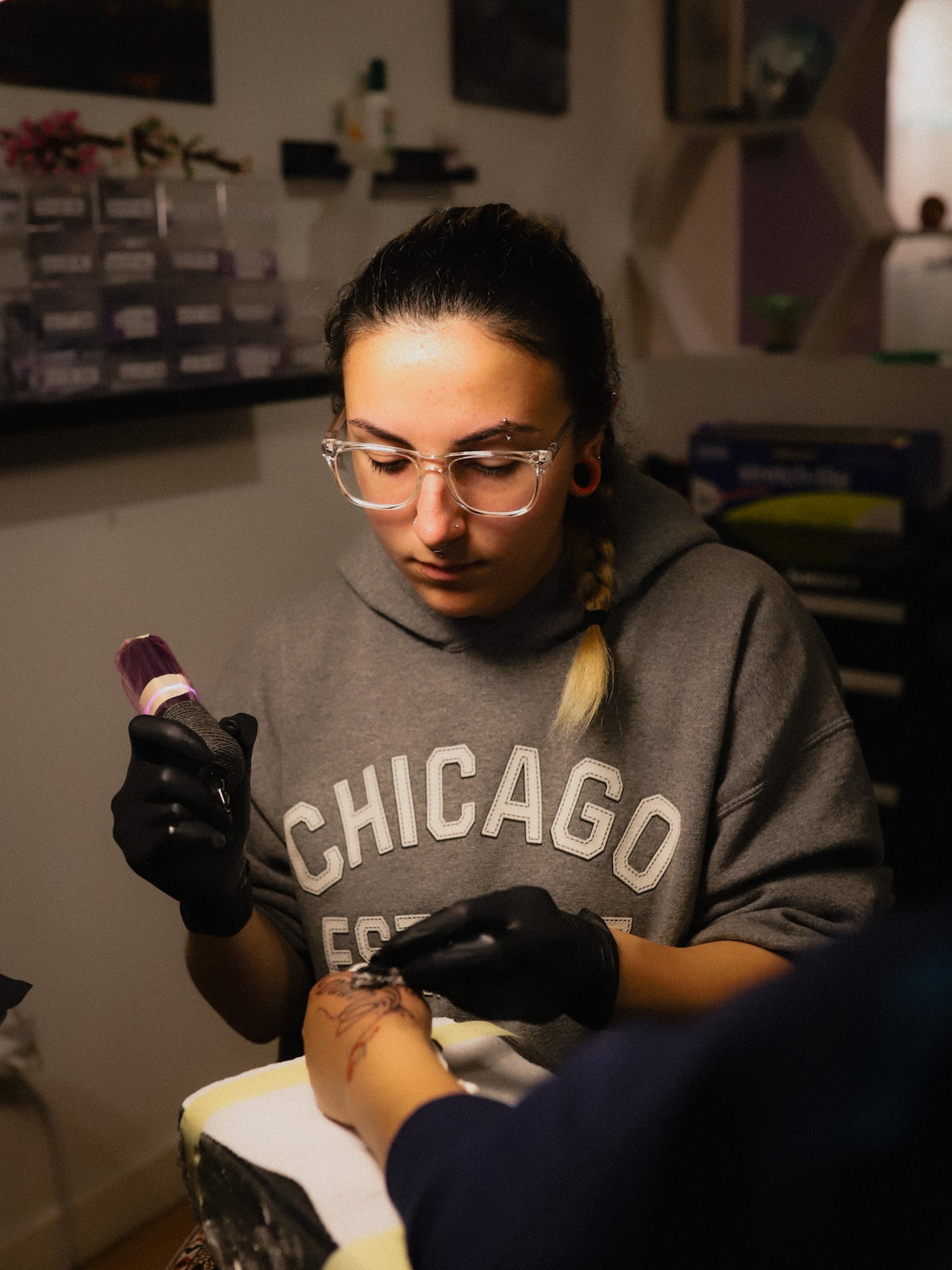 A woman wearing a gray Chicago hoodie and black gloves applies a tattoo to a client's hand in a tattoo studio.