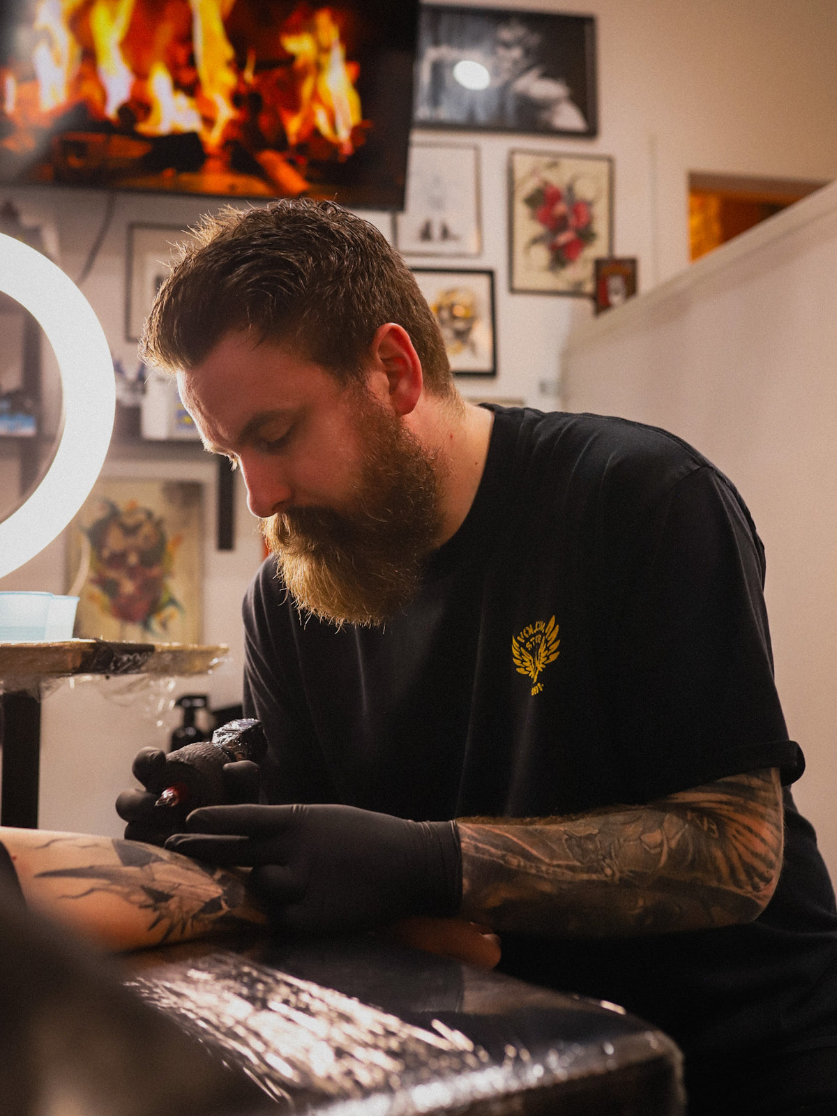 A bearded male tattoo artist wearing a black t-shirt with a yellow chest logo and black gloves, focused on tattooing a client's arm in a tattoo studio with a ring light and framed art on the wall.