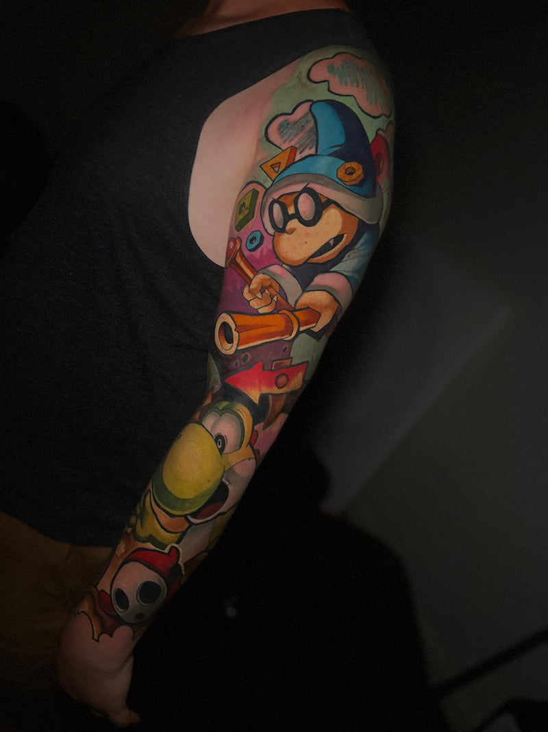 A close-up of a person's upper arm and shoulder featuring a colorful, full-sleeve cartoon tattoo done by Kelsie.
