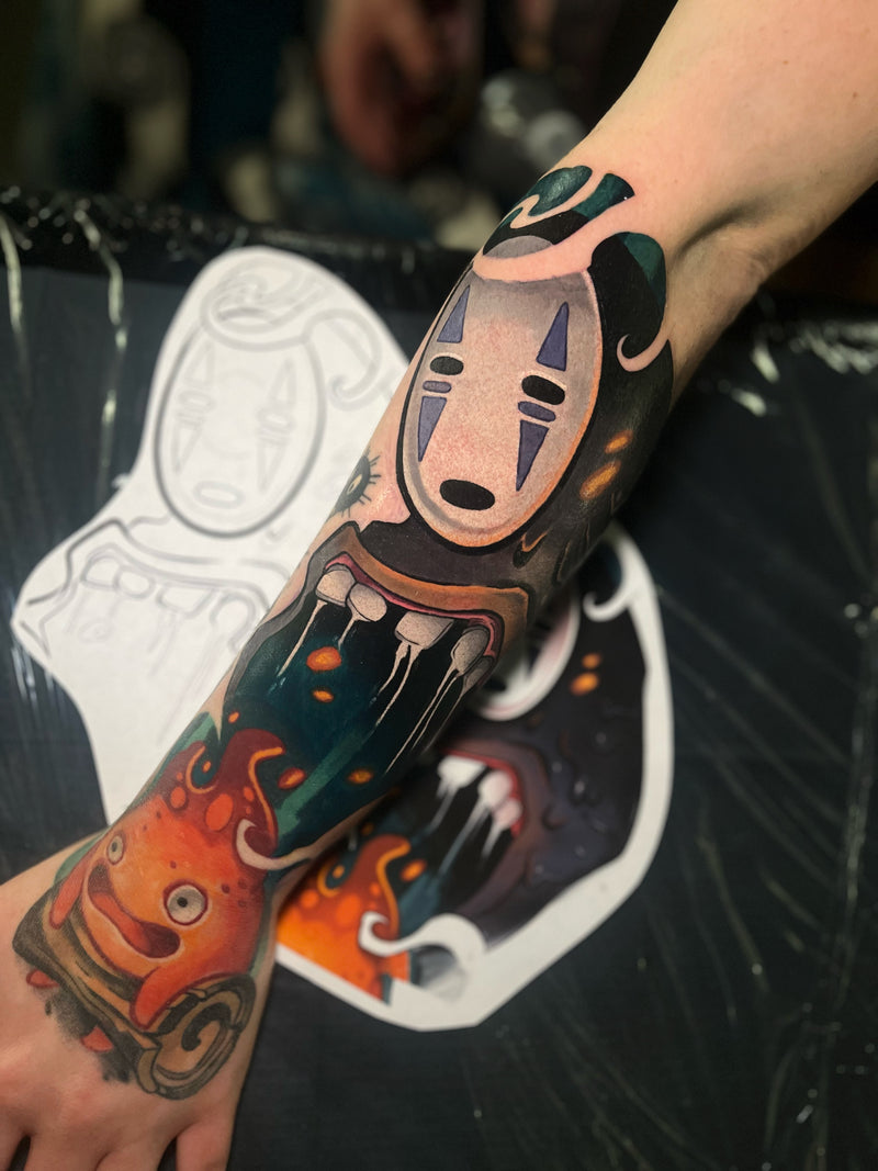 A close-up of a person's forearm and hand featuring a colorful, detailed tattoo  inspired by Studio Ghibi's animated films, including No-Face and the Little Mermaid done by Kelsie.