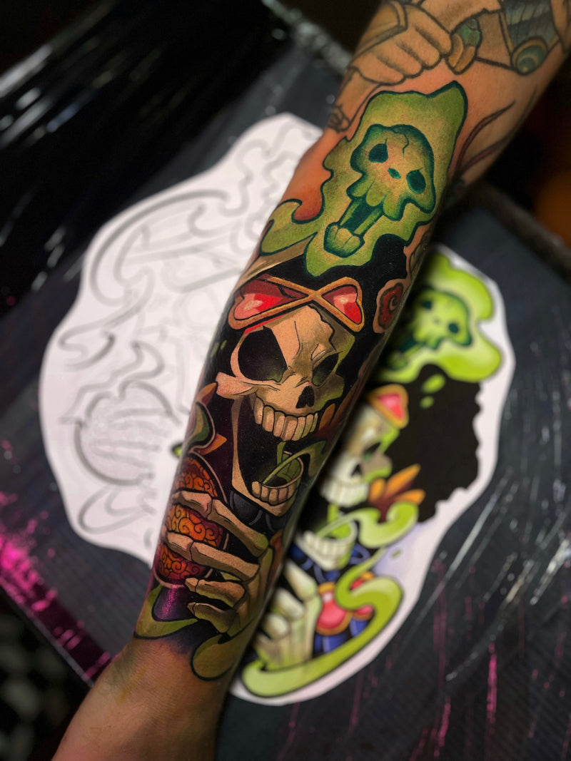 A close-up of a tattooed forearm featuring a colorful skull and ghost design in green, black, red, and orange, done by Kelsie