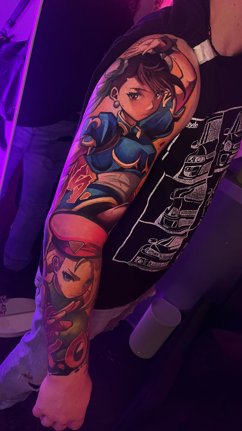 A close-up of a person's upper arm featuring a large, colorful sleeve tattoo with anime-style female characters done by Kelsie.