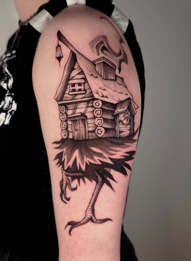A black and gray tattoo of a whimsical house with a long leg and claw-like foot, shown on a person's upper arm.