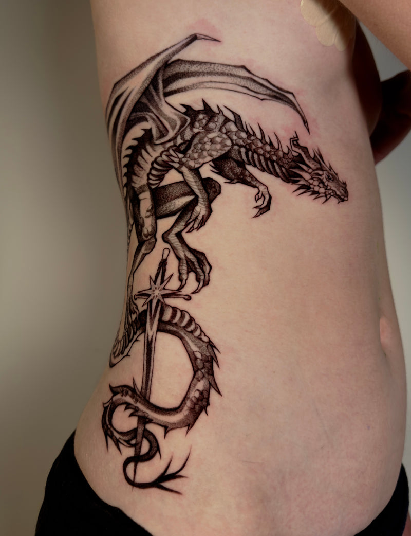 A detailed black and gray dragon tattoo on a person's side torso, showing the winged creature wrapping around a sword. Done by Mishel