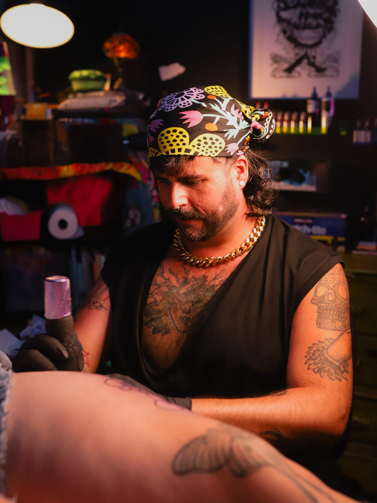A tattoo artist wearing a colorful patterned bandana, black sleeveless shirt, and gold chain necklace works on a client's tattoo in a dimly lit tattoo studio.